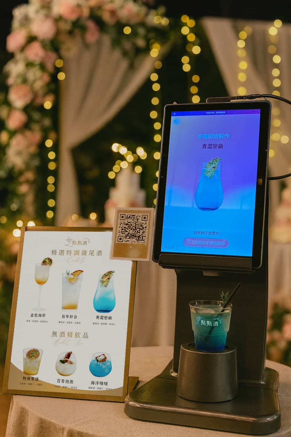 點點酒機台嵌在婚宴的花藝與燈串佈景中，旁邊立著酒單卡與掃碼點酒說明