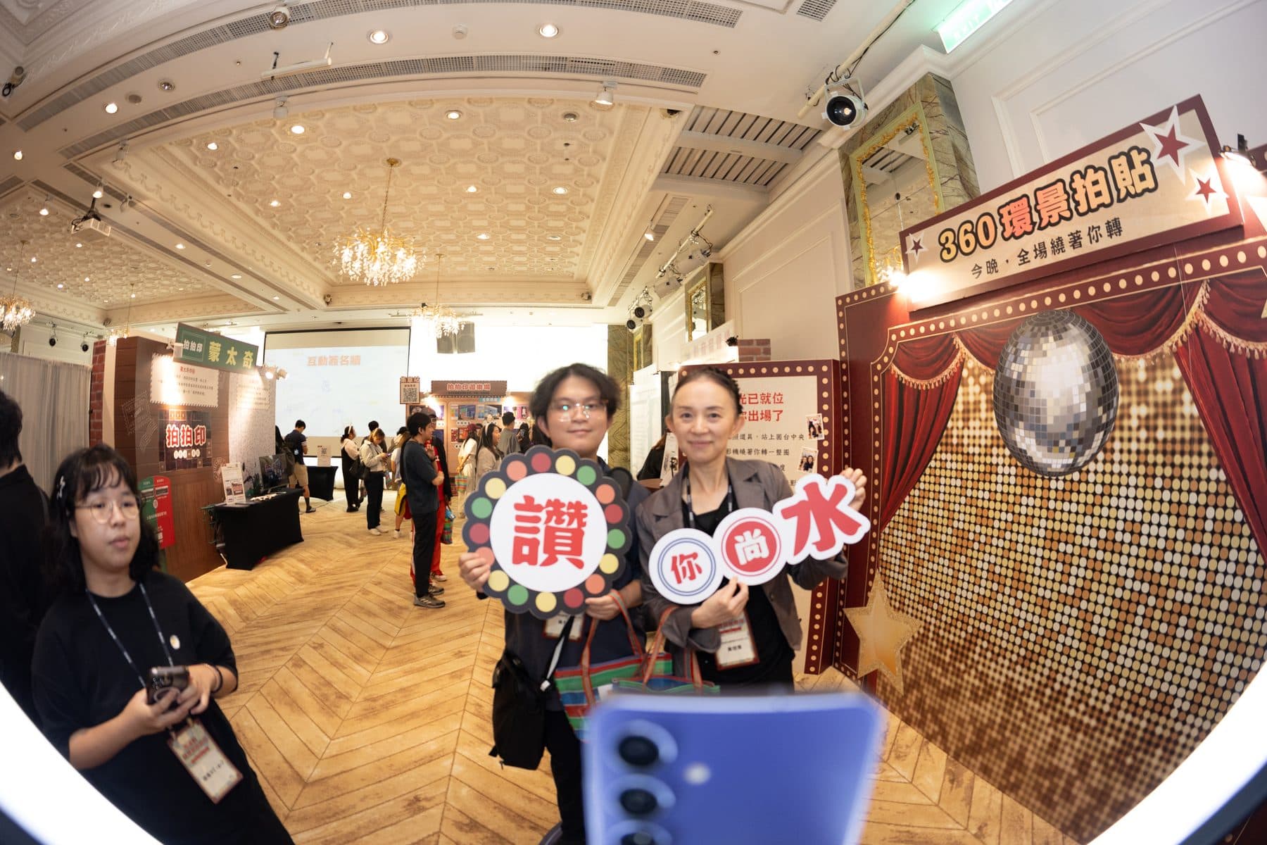兩位賓客手持「讚」與「你尚水」字樣手舉牌，在 360 環景拍貼背板前合影