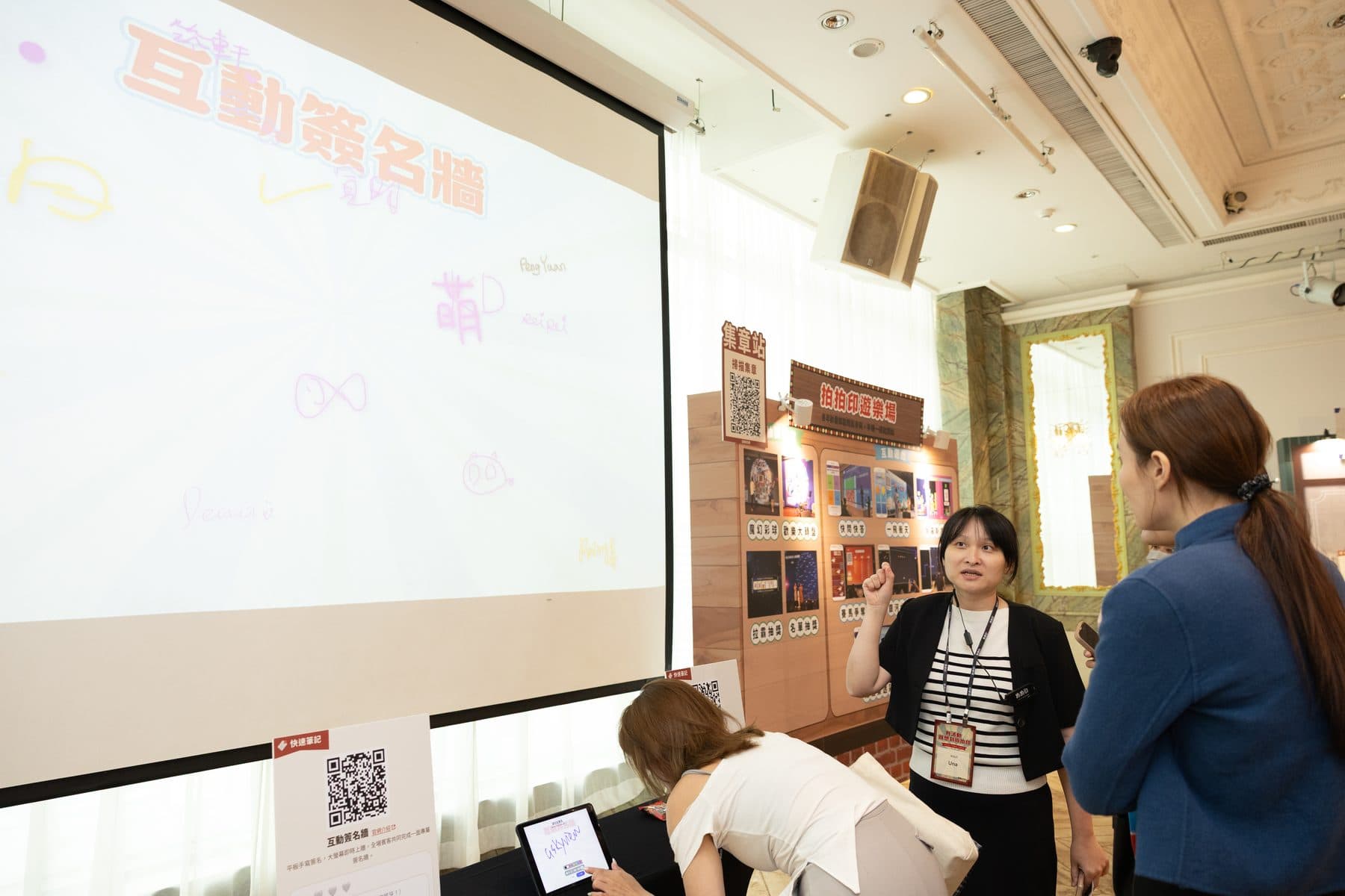 工作人員在投影布幕旁向賓客介紹互動簽名牆，幕上已有多筆手寫簽名
