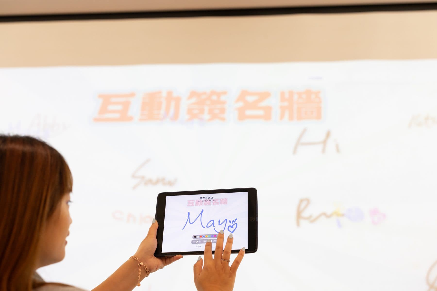 來賓在平板上手寫簽名「May」，後方布幕上已投影出互動簽名牆與其他人的簽名