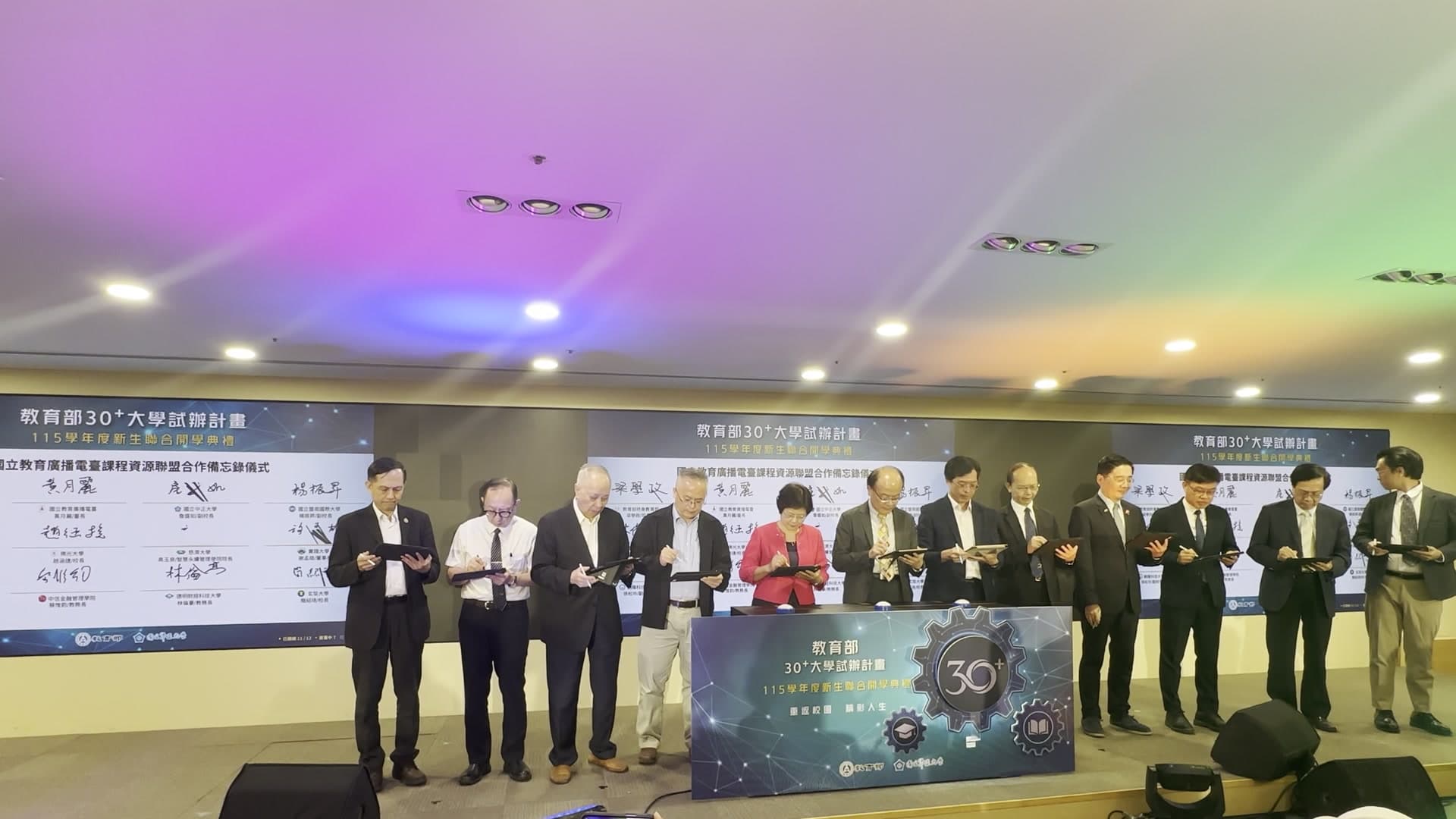 台下全景：12 位校長與長官在舞台大螢幕前同時在 iPad 上簽名